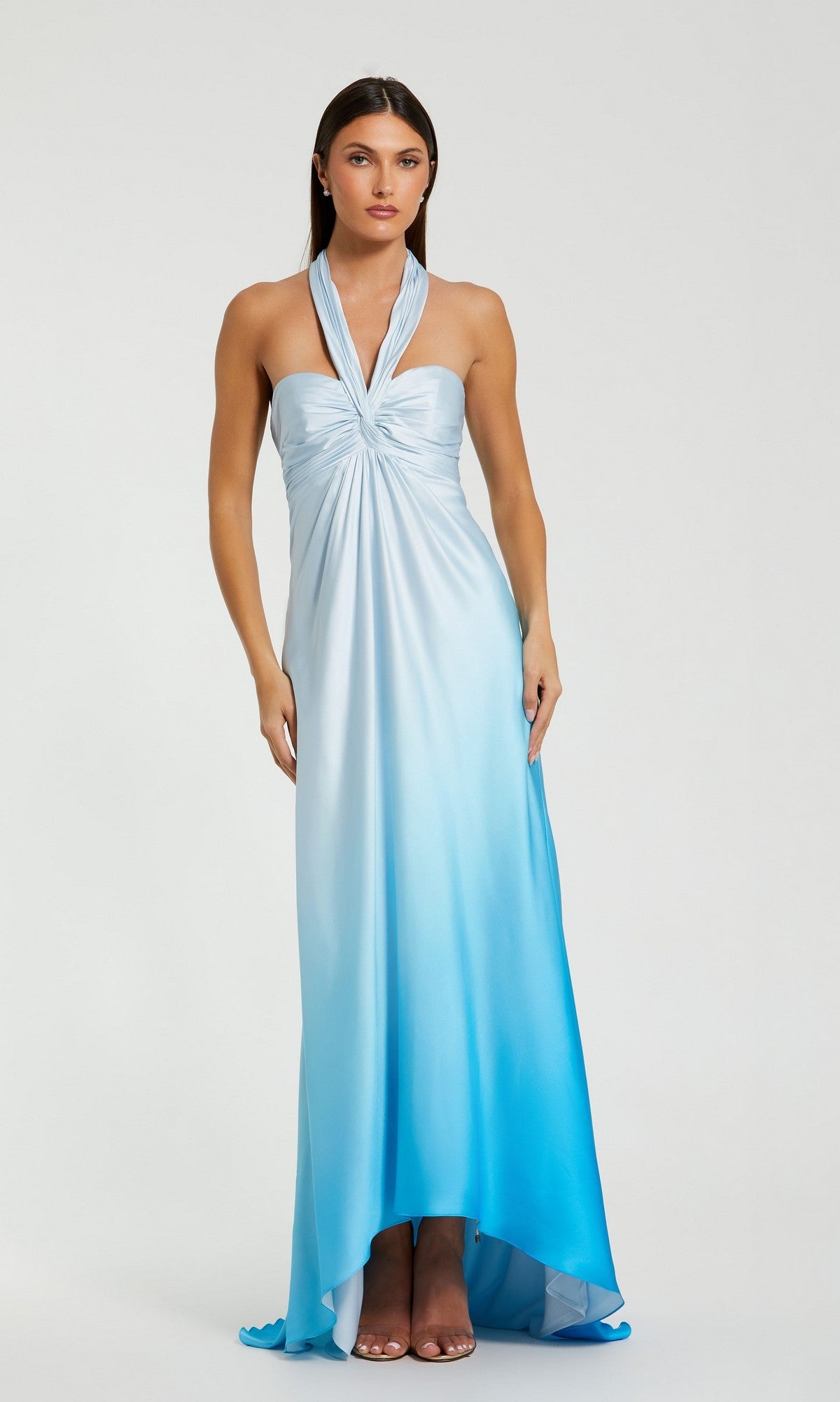 A woman in the Mac Duggal 68501 long formal dress, featuring a blue ombre charmeuse fabric, ruched bodice, halter sweetheart neckline, and subtle high-low hemline, stands against a plain light background.